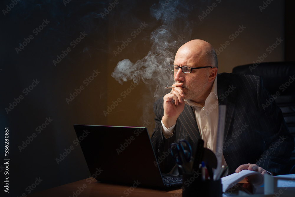 A man smokes while working. A man works in a dark office. A bald A man smokes while working. A man works in a dark office. A bald