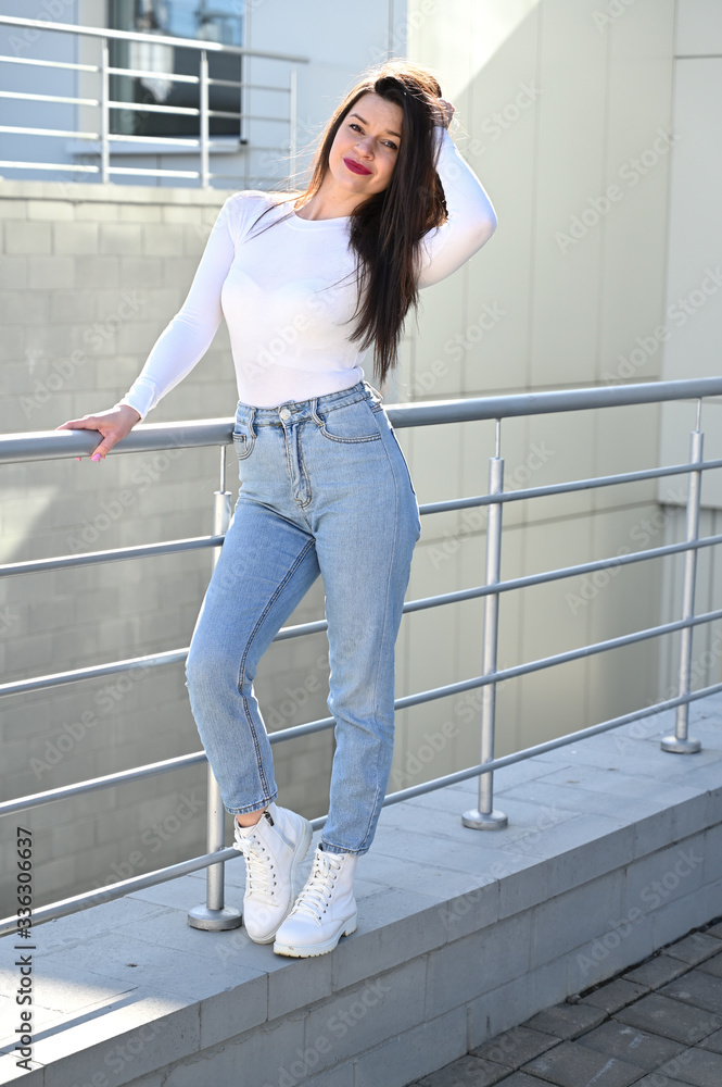 Photo of a caucasian brunette girl in a white blouse and blue jeans stands near the railing with a smile against the background of a gray wall of a building on a sunny spring day.