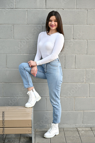 Vertical Caucasian portrait of a pretty brunette girl in a white blouse and blue jeans stands near the wall of a building on a spring day in the city. Model is happy to pose.