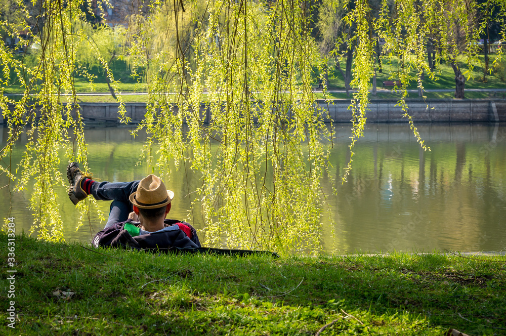 A man with a hat standing on the grass at the edge of a lake. Man enjoying a beautiful day in the park. A man with a hat standing on the grass at the edge of a lake. Man enjoying a beautiful day in the park.