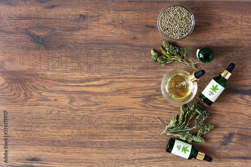 Green bottles of medical CBD oil with cannabis seeds and dried leaves top view on wooden background. Alternative medicine concept.