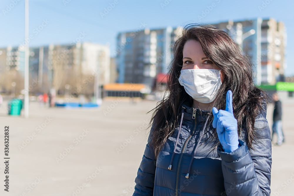 a beautiful girl in a medical anti virus mask warns by pointing her finger up while standing in a city square against the backdrop of shopping centers and a Playground