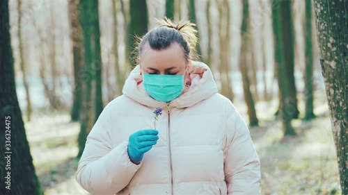 Woman in blue protective medical mask and gloves sniffing flower in forest through mask. Health care and medical concepts, coronavirus, virus protection. Close- up portrait. 4k photo