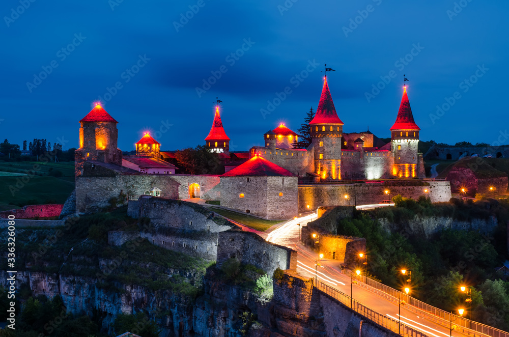 
Kamianets-Podilskyi Castle at night, illuminated by a red spire, a bridge with cars nearby