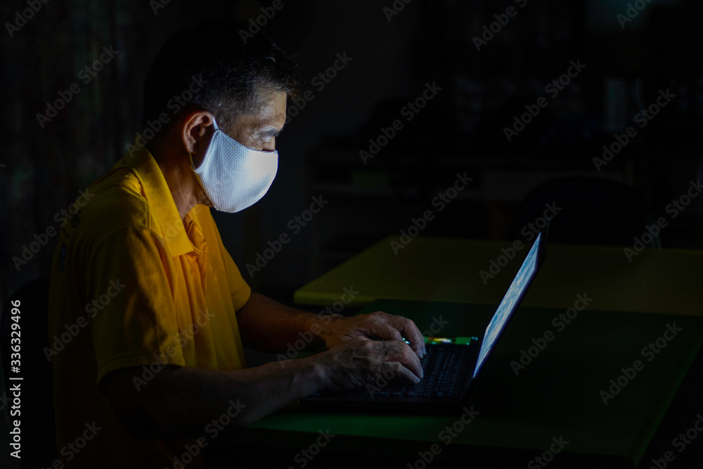 An Asian old business man uses his computer notebook while wearing face mask to connect with clients late at night from home. He keeps social distancing from members to prevent COVID-19 infection. An Asian old business man uses his computer notebook while wearing face mask to connect with clients late at night from home. He keeps social distancing from members to prevent COVID-19 infection.
