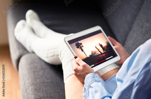 Woman watching tv series or movie stream with tablet on couch wearing cozy long socks. Girl streaming film with mobile device and on demand video (VOD) service. Digital entertainment site online.