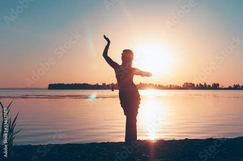 dark glowing silhouette of slim woman with hand up stay in dance pose near big river at sinrise