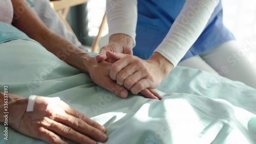 Unrecognizable friendly woman doctor sitting by patient in bed in hospital, holding hands.