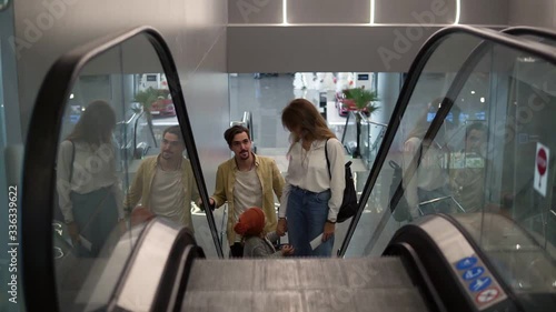 Young attractive caucasian family with suitcase going up by the escalator with their pretty daughter hold by mothers hand at the airport. Happy family vacation together