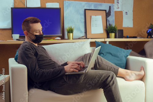 A man in a black mask lies on a sofa and works at a computer. Quarantine, masks, work at home.