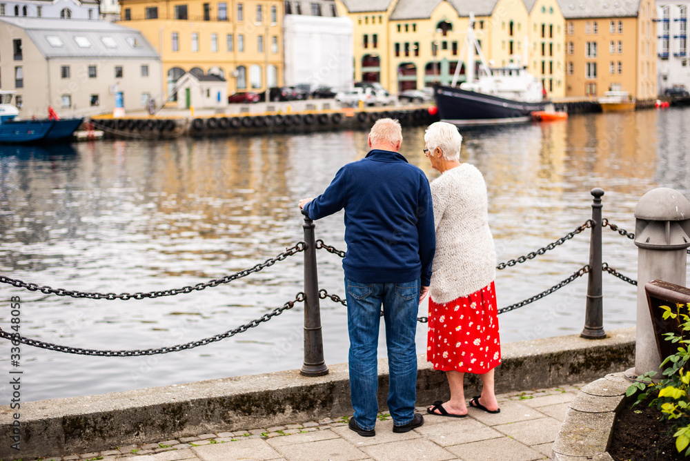 Fototapeta premium Elderly couple standing at chain fence next to water edge in the city. Watching town life during walk