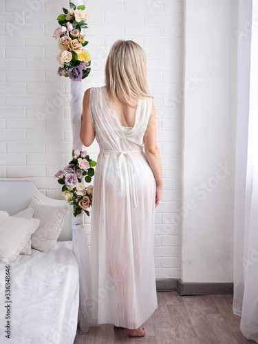 Three-quarter portrait of young pretty blond woman bride, wearing white lace bridal peignoir, standing in the light room with white furniture, with her back to the camera, on the wedding morning