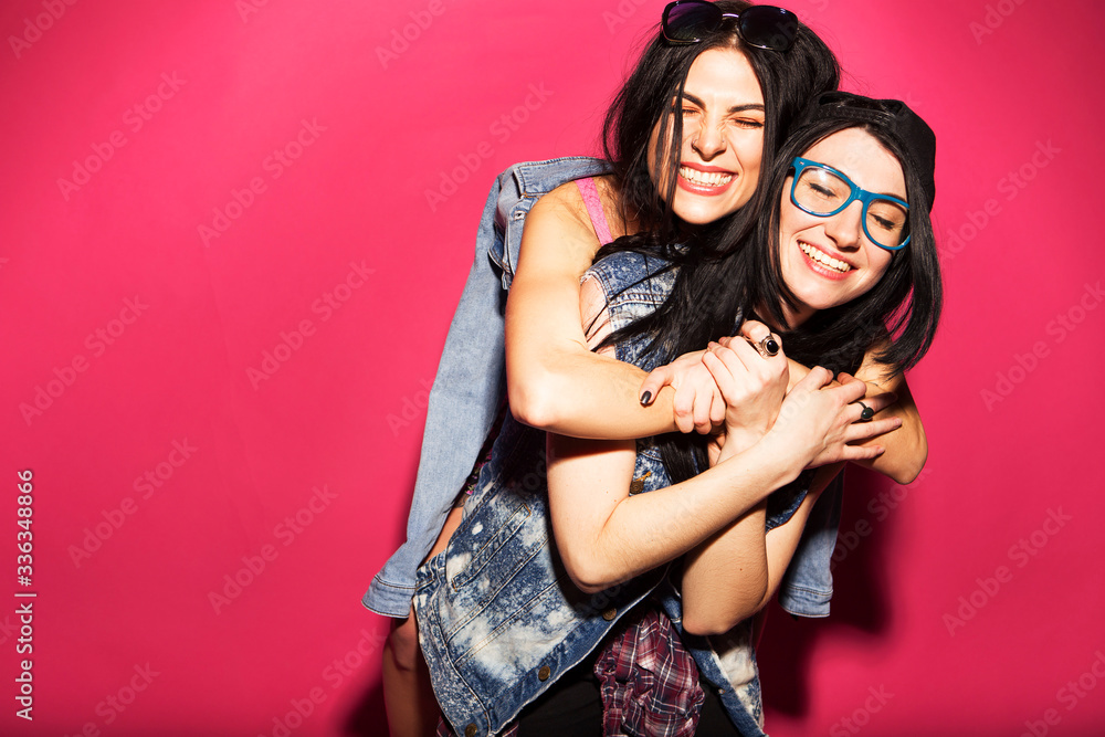 Two caucasian brunette hipster woman in casual stylish jeans outfit having fun chewing bubblegum and holding skateboard. They standing on a bright pink background. Cheerful, happy emotions Two caucasian brunette hipster woman in casual stylish jeans outfit having fun chewing bubblegum and holding skateboard. They standing on a bright pink background. Cheerful, happy emotions