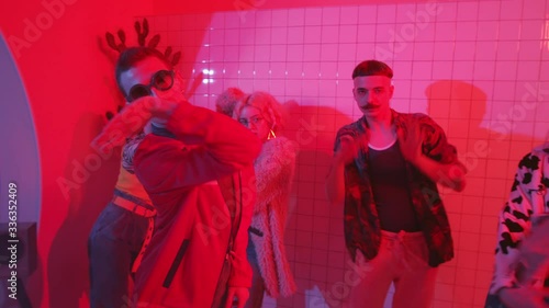 Young eccentric man in trendy wear and sunglasses dancing expressive vogue with group of stylish friends in room with color neon light and fog photo