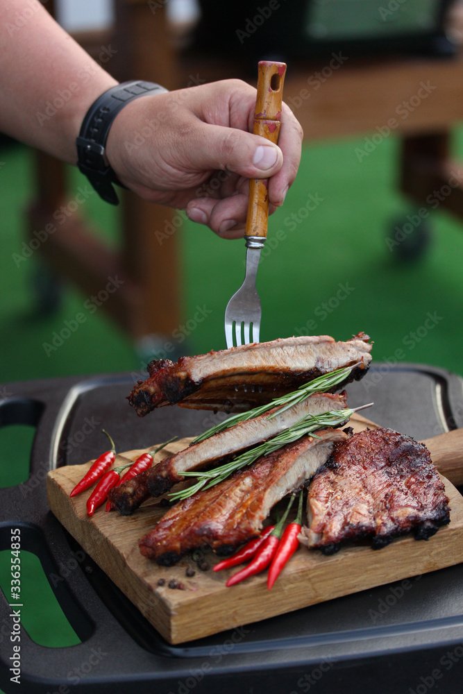 Fried pork ribs with a fried crust with spices, rosemary and chili pepper on a wooden board on the outdoor  with fork and hand. Meat on grill. Background image, copy space