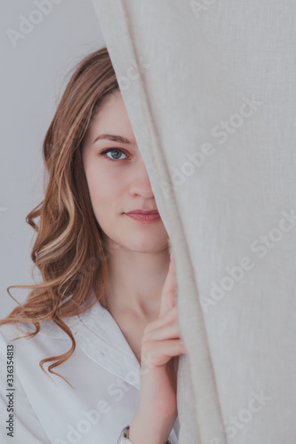 a young pretty woman looks out from behind white curtains. half face hidden behind a curtain © Марина Кудревич
