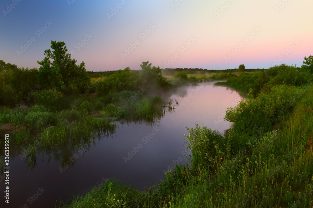 River in the early morning at dawn. Delicate dawn sky and fog rising above the water, lush greenery on the banks. Summer spring wild landscape by the river. Selective soft focus.