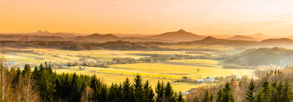 Fototapeta premium Hilly landscape illuminated by evening sunset. Green grass fields and hills on the horizont. Vivid spring rural countryside. Ralsko Mountain, Czech Republic
