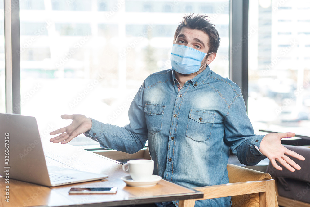 Not sure! Portrait of confused young man with medical mask in blue shirt sitting, working on laptop with raised arms and puzzled face, looking at camera. indoor working and health care concept. - obrazy, fototapety, plakaty 