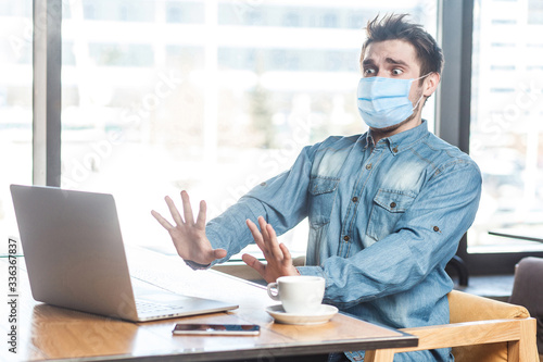 Portrait of scared young businessman with surgical medical mask in blue shirt sitting, shocked looking at laptop display with raised arms and stressed face. Indoor working and health care concept.