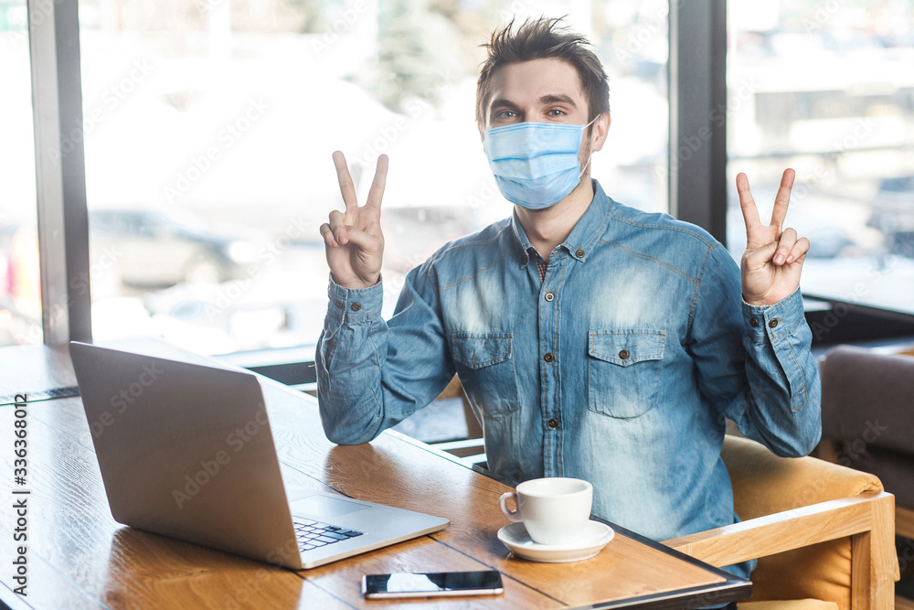 Φωτογραφία Portrait of handsome successful positive young man with surgical medical mask in blue shirt sitting and looking at camera with victory gesture