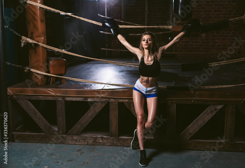Young girl in sportswear: a t-shirt and shorts wearing boxing gloves. Athletic woman posing on a background of a boxing ring in the dark gym. Power and motivation concept