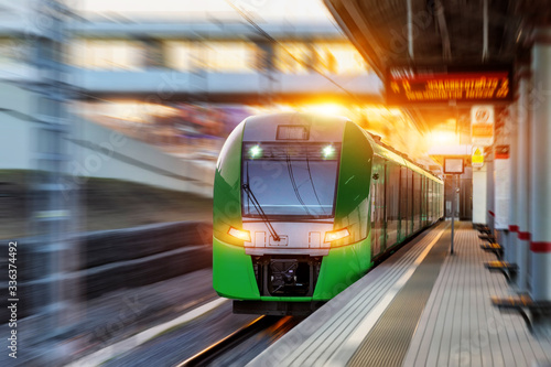 Passenger electric train arrives at the station in urban landscape at sunset evening time rush hour.