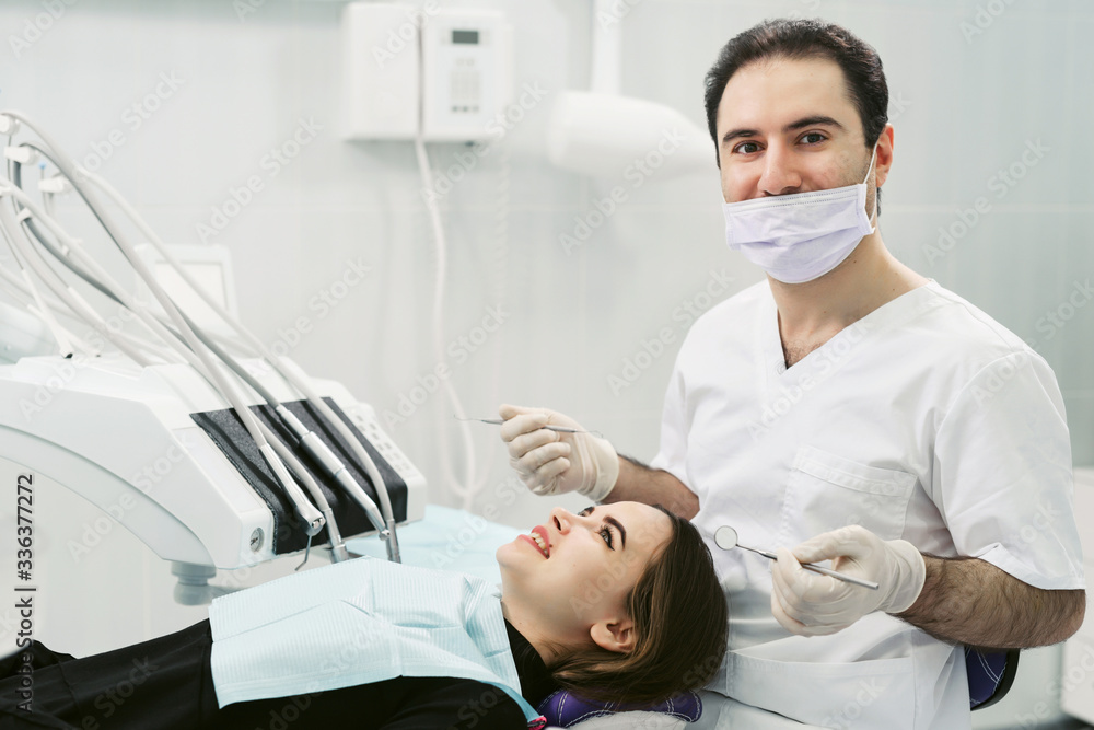 Portrait of a friendly smiling male dentist with patient in the office of a modern dental clinic. Doctor in disposable medical facial mask. Portrait of a friendly smiling male dentist with patient in the office of a modern dental clinic. Doctor in disposable medical facial mask.