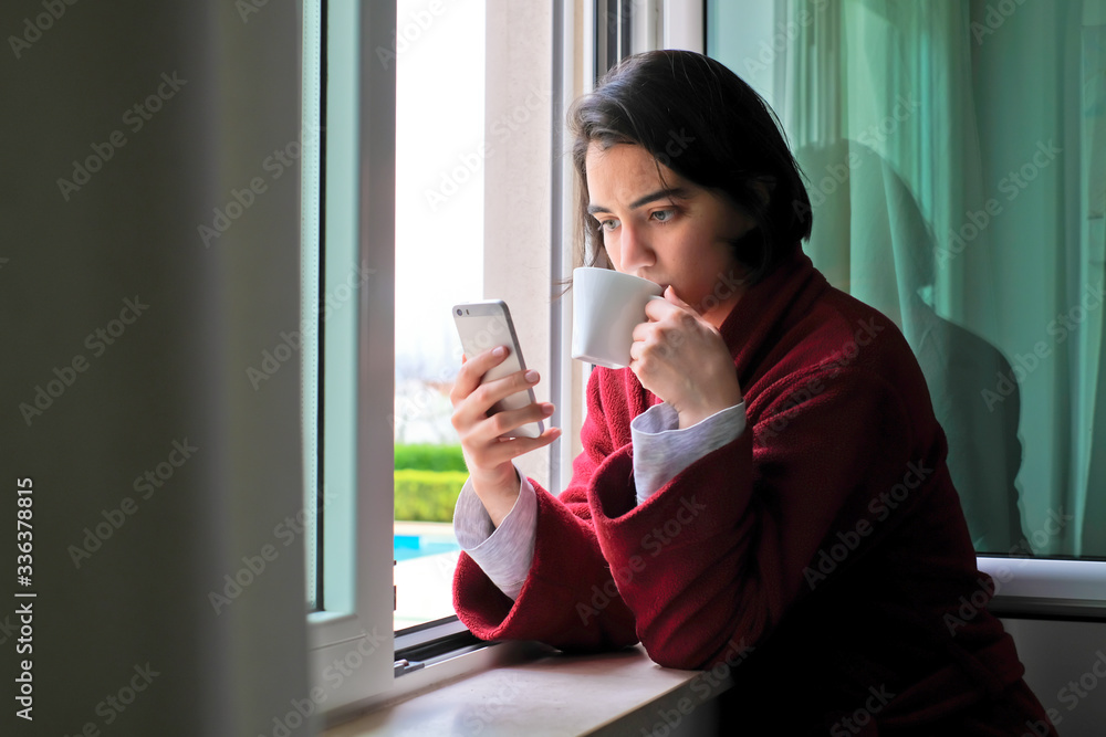 A caucasian woman reading the news on her phone while drinking a coffee by the window wearing pajamas and a red robe at home during self isolation A caucasian woman reading the news on her phone while drinking a coffee by the window wearing pajamas and a red robe at home during self isolation