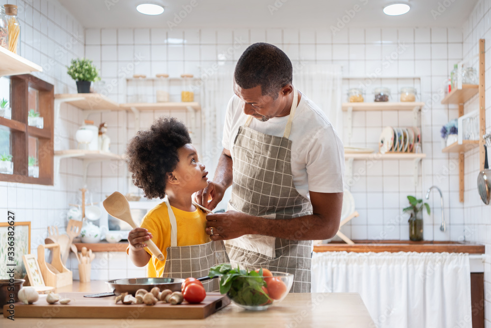 Happy family with African father and son dress up together before cooking in the white kitchen. Single Dad Chef with black kid helper in yellow shirt preparing food and looking at Each Other at home. Happy family with African father and son dress up together before cooking in the white kitchen. Single Dad Chef with black kid helper in yellow shirt preparing food and looking at Each Other at home.