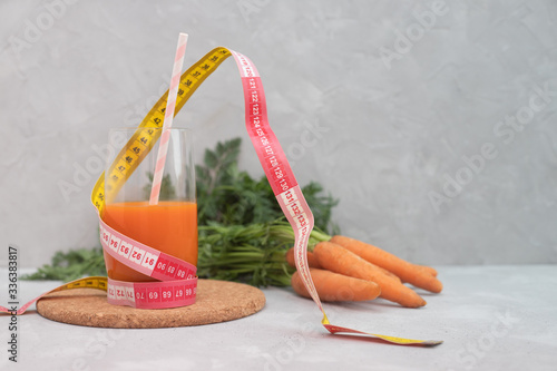 A glass with fresh carrot juice with a straw and around it a measuring tape, centimeter. In the background a bunch of carrots with green tops. Dietary nutrition