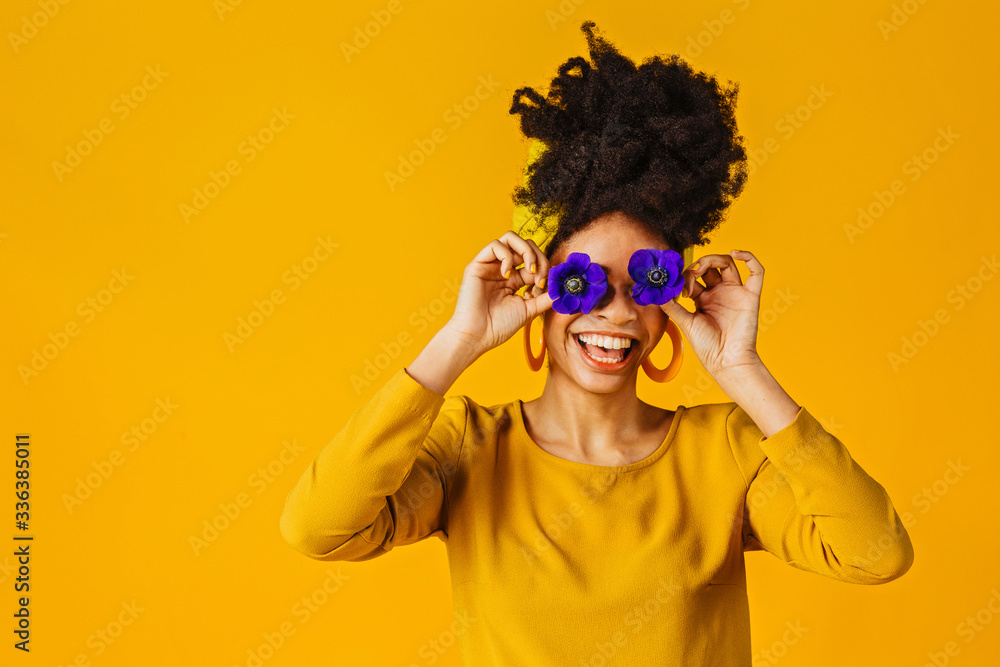 Portrait of a happy young woman holding two flowers over eyes, laughing and having fun, against yellow background Portrait of a happy young woman holding two flowers over eyes, laughing and having fun, against yellow background