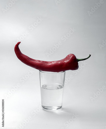 A glass cup half full of water stands on a light background. Red pepper lies on a glass. Close-up