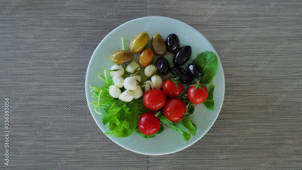 Top view, cherry tomatoes, black and green olives and mozzarella cheese balls. Arugula salad to the edge. The plate rotates. Healthy food concept. The plate is spinning on the tablecloth. Top view, cherry tomatoes, black and green olives and mozzarella cheese balls. Arugula salad to the edge. The plate rotates. Healthy food concept. The plate is spinning on the tablecloth.