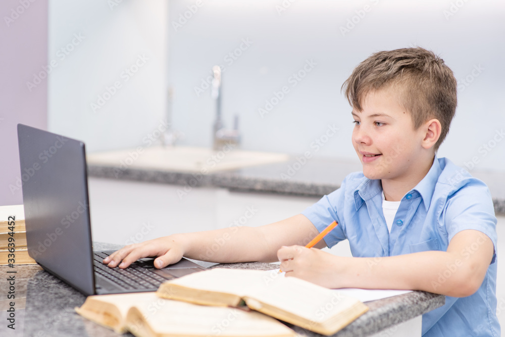 A boy joyfully looking at the screen engaged in distance learning at home using a laptop writing down his homework in a notebook. Distance Learning, Home-based Learning A boy joyfully looking at the screen engaged in distance learning at home using a laptop writing down his homework in a notebook. Distance Learning, Home-based Learning