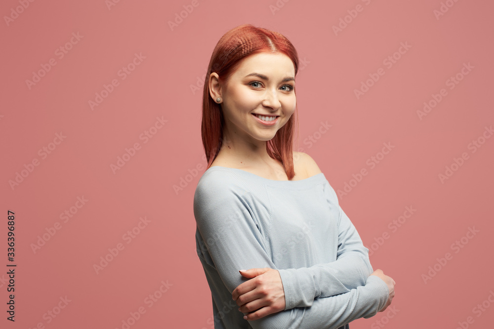 Obraz premium Smiling ginger girl in gray sweater with arms crossed standing in studio isolated on pink background