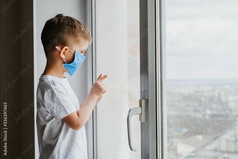 a child in a medical mask is sitting at home in quarantine because of coronavirus and covid -19 and looks out the window.