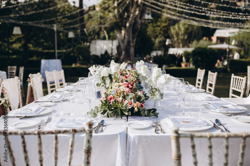 Wedding banquet. Tables in a green meadow are decorated with flower arrangements, on the tables are white tablecloths, plates with napkins, glasses and candles, cutlery. White bulbs hang over tables © AlexGukalovUkraine