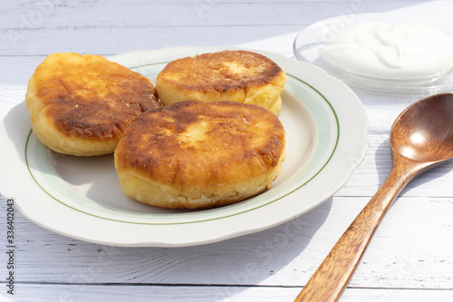 Three fried curd cheesecakes with a wooden spoon and sour cream on a transparent saucer on a white plate on a plank table. Delicious Breakfast of flour, eggs and cottage cheese