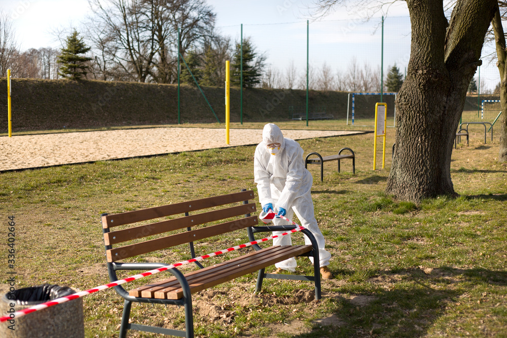Osoba ubrana w kombinezon ochronny zabezpiecza ławki w parku by uniemożliwić korzystanie z nich w czasie pandemii - obrazy, fototapety, plakaty 