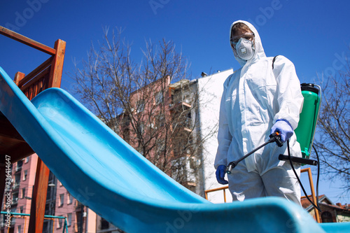 Children playground disinfecting and sanitizing against virus and disease. Man in white protective suit spraying disinfectant on slide to stop spreading coronavirus or COVID-19. photo