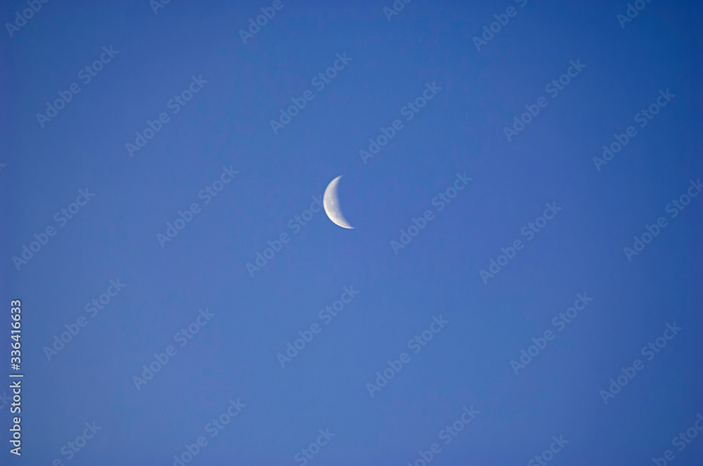 A slim sliver of the silvery moon set on an expanse of clear and cloudless blue sky.