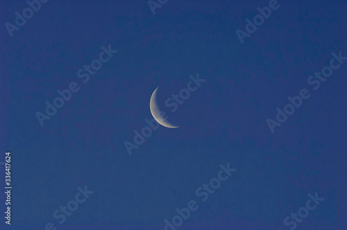 A slim sliver of the silvery moon set on an expanse of clear and cloudless blue sky.
