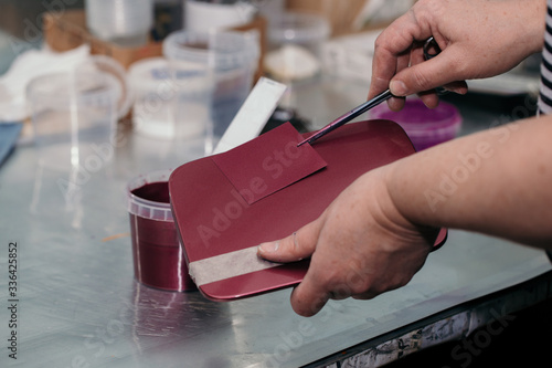 The painter’s hands hold a test plate with a paint applied on it, matched to the color of the car’s hatch and the burgundy car part