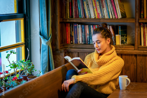 On the couch in front of the window, a woman caught up in reading. The woman at the table drinking coffee and reading a book
