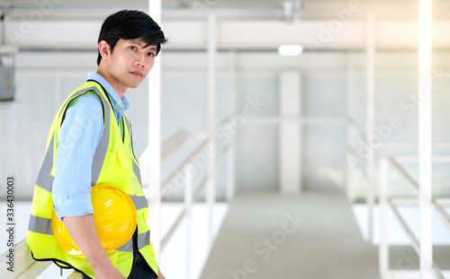 Junior engineer standing in the plant holding helmet and looking outside with copy space. Wearing safety vest. Feeling happy and smile on work. Engineering and design concept.