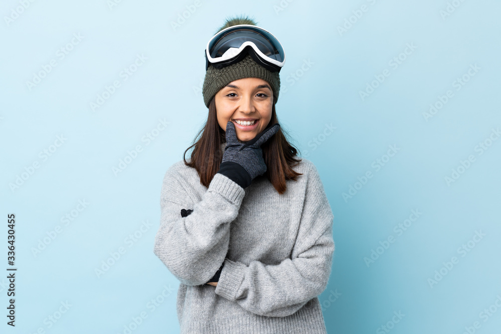 Mixed race skier girl with snowboarding glasses over isolated blue background happy and smiling. Mixed race skier girl with snowboarding glasses over isolated blue background happy and smiling.