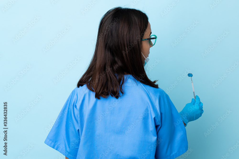 Young brunette mixed race dentist woman holding tools over isolated background in back position and looking side Young brunette mixed race dentist woman holding tools over isolated background in back position and looking side