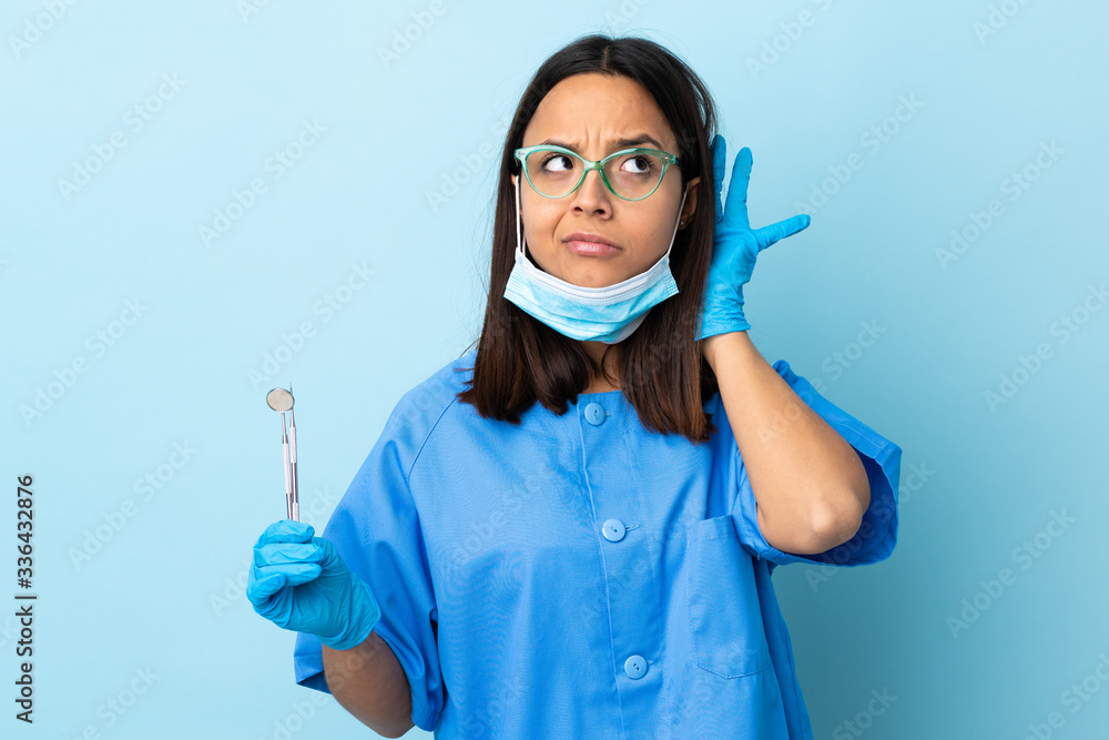Young brunette mixed race dentist woman holding tools over isolated background listening to something by putting hand on the ear Young brunette mixed race dentist woman holding tools over isolated background listening to something by putting hand on the ear