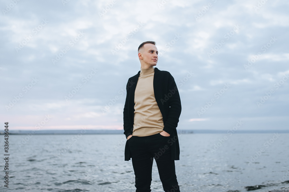 Stylish man, the groom in a black cardigan stands on the pier, against the background of the sea, river and sky with clouds. Photography, concept. Stylish man, the groom in a black cardigan stands on the pier, against the background of the sea, river and sky with clouds. Photography, concept.